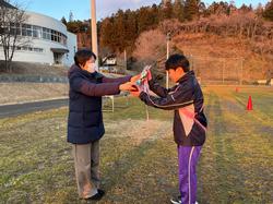 芝生の運動場でジャージを着た男子生徒が女性から優勝トロフィーを受け取っている写真