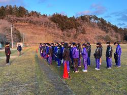 芝生の運動場にジャージを着たたくさんの生徒たちが整列し、前に立った人物の話を聞いている様子の写真