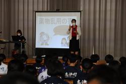 資料を映したスクリーンの前で講師の女性が話し、椅子に座った生徒たちが講話を聞いている様子の写真