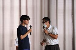 男子生徒とサングラスとマスクを身に着けた男性がそれぞれマイクを持ち、誘惑を断る練習をしている様子の写真