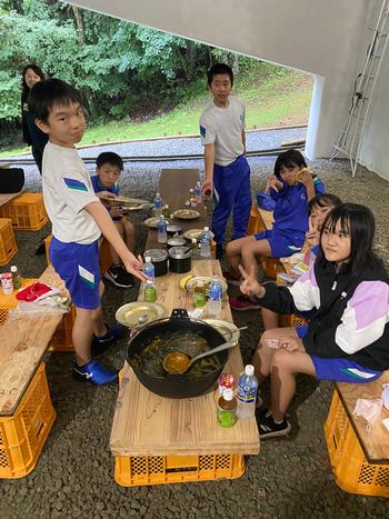 空になった大きな鍋を指さして立っている男の子2名と、その周りに座ってピースをしている女の子たちの写真