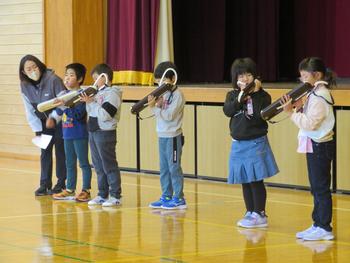 5名の児童が舞台の前に1列に並び、先生に見守られながら鍵盤ハーニカの演奏をしている写真
