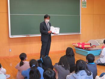 黒板の前で男性教諭が絵本を開きながら両手で持ち、床に座っている子供たちに読み聞かせを行っている写真