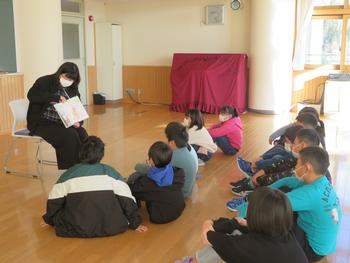 床に座った子供たちが前方の女性の読み聞かせに耳を傾けている写真