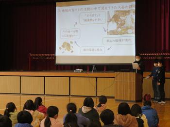 舞台の大きなスクリーンに映し出された内容を演台に立っている児童が発表をしている写真