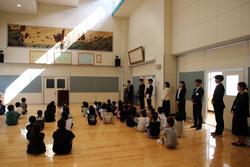 広い室内の右側に先生方が並んで立ち、室内中央付近に生徒たちが整列して床に座っている始業式の様子の写真