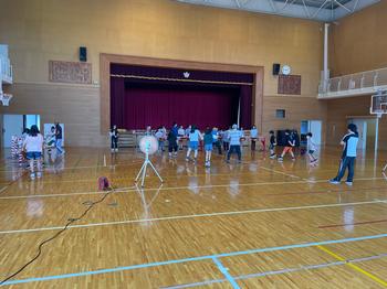 大型扇風機が複数設置された体育館内で舞台の前に立って歌の練習や合奏の隊形確認をしている児童たちの写真