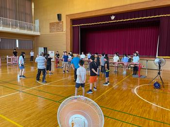 大型扇風機が複数設置された体育館内の舞台上には鍵盤ハーモニカなどの楽器を持った児童が並んでおり、その前で児童たちが円形や列を作って立っている様子を写した写真