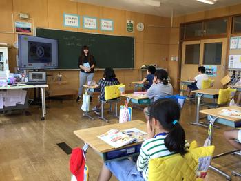 教室の黒板前に女性のALTが立ち、席に着いた生徒たちに向かって、左に置いたモニターを使いながら授業を行っている様子の写真