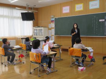 黒板の前に女性教諭が立ち子供たちが机に座って授業を受けている様子を写した写真