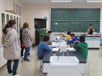 理科室で各机に座った生徒の周りに保護者の方が立って授業の参観をしている写真