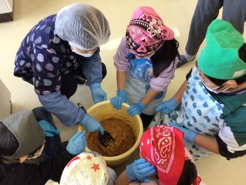 真ん中にバケツがあり入っている味噌を講師の先生がまぜている様子をエプロン姿の子供が覗き込んでいる様子を上から写した写真