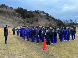 芝生の運動場でジャージ姿の生徒たちが整列し、向かい合って立った人物の話を聞いている写真