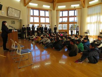 小学生たちが床に整列して座り、演台に立つ女性の話を静かに聞き、後方には教職員が見守っている全校集会の写真