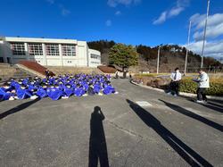 校舎の手前の舗装された広場に、ジャージ姿の多くの生徒たちが整列して座り、その前に2人の男性が立っており、手前には3人の人物の影が地面に長く伸びている写真