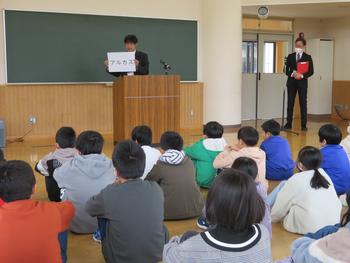 児童たちが床に座って、前方の演壇（教壇）で「アルカス」と書かれた紙を掲げて話をしている人物の話を聞いている写真