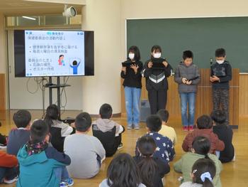 黒板の前に4名の生徒が並びモニターを使いながら、床に座ったたくさんの生徒の前で発表を行っている写真