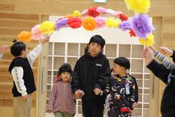 色とりどりの紙の花で装飾されたアーチの下を、男の子と女の子が手を繋いで通っている写真