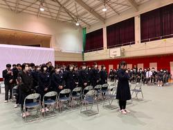 体育館に集まった制服姿の生徒たちが椅子の前で姿勢を正して立ち、前に立った1人の女子生徒が話をしている写真