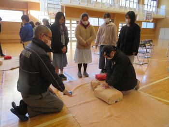 体育館で、南三陸消防署の男性の指導の下、1人の女子児童が人形を使って心肺蘇生法の実践をし、横に立った4人の女子児童が見学している様子の写真