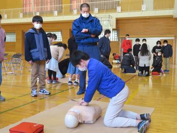 体育館でグループに分かれて心肺蘇生法の練習をする中、手前のグループの男子児童が人形の胸に手を当てて実践練習をし、周りで南三陸消防署の男性と男子児童が見守っている様子の写真