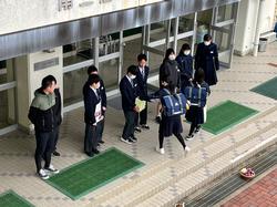 校舎の昇降口で横一列に並んだ生徒たちが募金活動を行っており、前を通っている2人の女子生徒が募金を行っている様子の写真