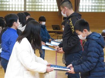 体育館で複数の在校生が、向かい合って立った6年生たちに自作のプレゼントを手渡している様子の写真