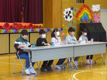 カラフルな飾り付けがされた体育館のステージ前で、それぞれの前に置かれたコップの横に手を置き、4人の児童がテーブルに並んで座っている写真
