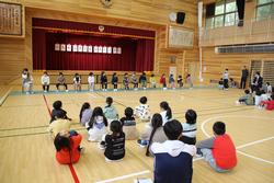 ステージ前の並べられた椅子に座っている生徒、体育館の床に座っている生徒たちがいる1年生を迎える会全体を写した写真