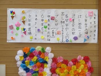 木目調の壁に貼られた、6年生を送る会の手書きの式次第と、その下にカラフルな花の飾り付けが施されている様子の写真