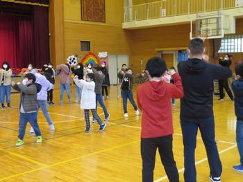 体育館で、大勢の児童と先生方が一緒に、円を描きながら腕を上げて踊っている様子の写真