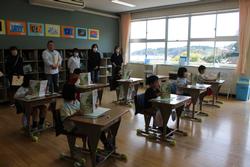 教室内で、複数の小学生たちが自分の机で真剣に教科書を読み、その後ろでは保護者たちが静かに見守っている様子の写真