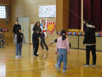 体育館のステージ前で、複数の小学生と先生が縄跳びを飛んでいる様子の写真