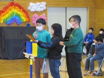 体育館で、虹色の装飾を背景に、マイクを持った男子児童が、他の児童たちと協力し6年生に向けて話をしている様子の写真