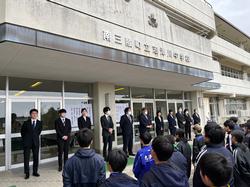 校舎の昇降口前に集まって整列した生徒たちの前に、スーツを着た11人の職員が並んで立っている写真