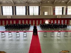 体育館に十字に敷かれた赤い絨毯の中央あたりに女性が立っており、その前には左右に並んだ椅子に大勢の生徒たちが女性の方を向いて座っている写真