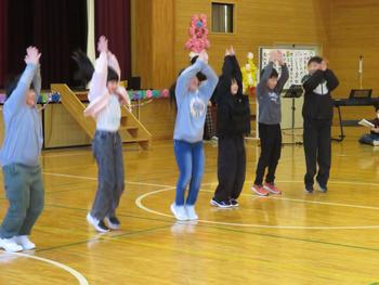 体育館の舞台の前で6人の児童が元気よくジャンプしながら踊っている「六年生を送る会」の一場面の写真