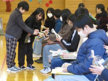左側の児童が立って、座っている六年生にプレゼントを手渡し、六年生たちは手にプレゼントやメッセージカードを持ち、嬉しそうに眺めている写真