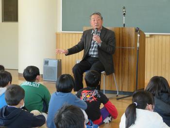 教室の黒板の前で、阿部さんが椅子に座ってマイクで話し、それを床に座った児童たちが聞いている様子の写真