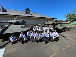 屋外に展示された戦車の前で、多くの学生が制服姿で並び記念撮影をしている写真