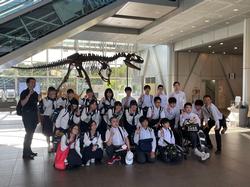 館内に展示された大きな恐竜の骨格標本を背にして、多くの学生が制服姿で集まり記念撮影をしている写真