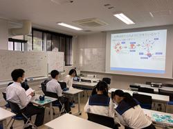 教室内で制服姿の学生たちが机に座り、前方のスクリーンに映し出された資料を見ている写真