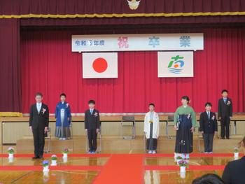 体育館の舞台前に設置したひな壇に、袴やスーツを身につけた児童と教員たちが整列して立っている様子の写真