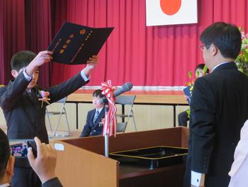 演台の前に立った男子児童が先生から受け取った卒業証書を高く掲げている様子を捉えた写真
