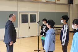 スーツ姿の男性が複数の子供たちと向き合い、マイクを前に何かを話している様子の写真