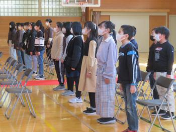 前後2列に並んだ椅子の席の前で、たくさんの在校生たちが姿勢を正して立っている写真