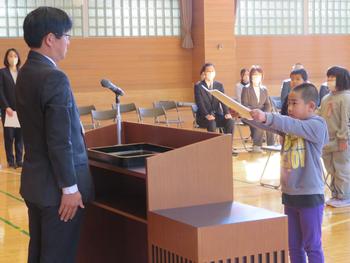 体育館の壇上で、小学校の児童が修了証書を先生から受け取る様子の写真