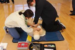 床に置かれた心肺蘇生法の練習用人形を使い、2名の女性教師が協力して胸骨圧迫の訓練をしている様子の写真