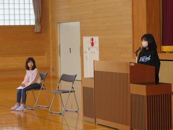 体育館の壇上の前で、一人の女子児童がマイクを使って発表し、もう一人の児童が椅子に座って話を聞いている様子の写真
