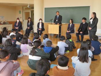 床に座って真剣に話を聞いている子供達の前で、黒板前に並んだ新しい先生型が並んで1人の先生がマイクを使って話している写真